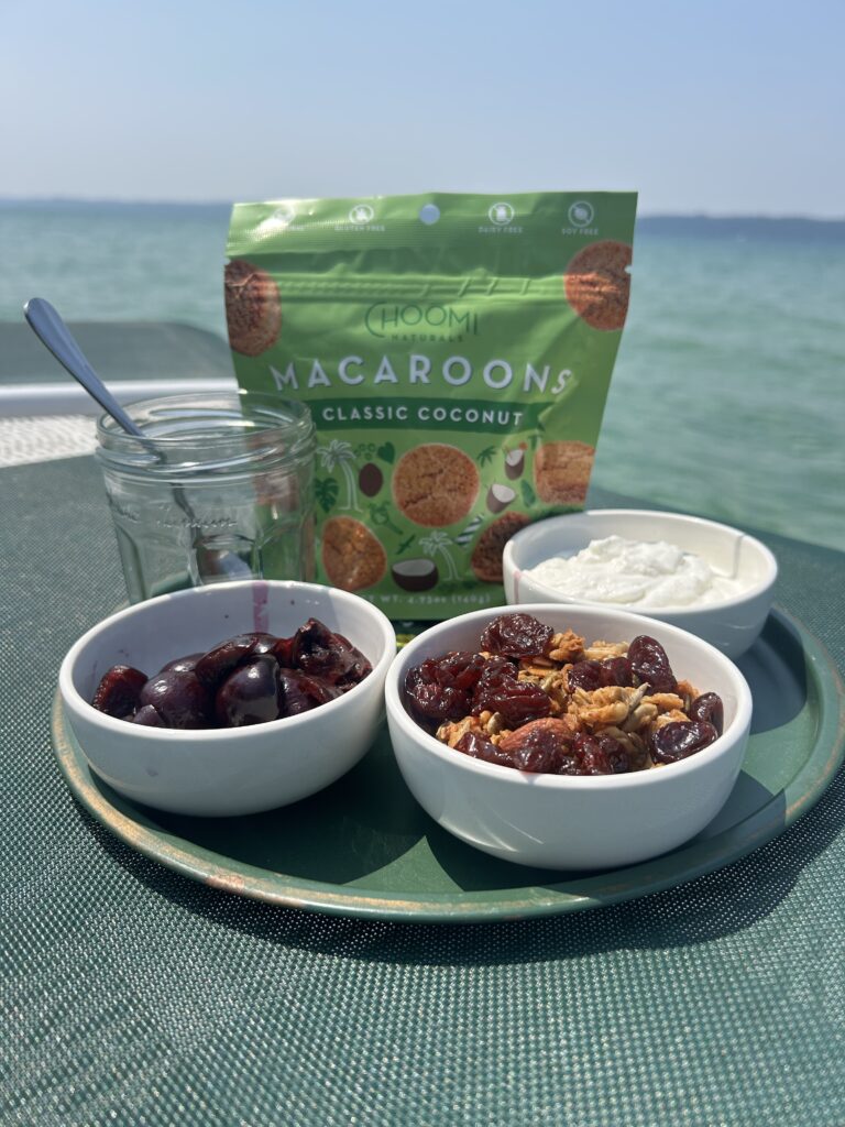 A top-down shot of a green tray with bowls of cherries, a dried fruit and nut mix, and a white creamy substance. A bag of "CHOOMI MACAROONS CLASSIC COCONUT" is in the background, with a glass jar and spoon.