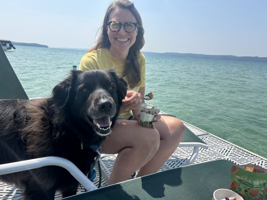 A person in glasses and a yellow shirt smiles at the camera, holding a jar of food, with a black dog next to them. They are sitting on a boat with water stretching behind them.